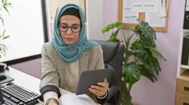 Ofis masasında bilgisayar ve bitkilerle tablet kullanan tesettürlü olgun bir kadın.