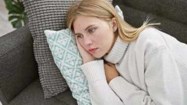 Beyaz kazak giyen, sarı saçlı, mavi gözlü, beyaz saçlı beyaz bir kadın, kapalı bir kanepedeki yastıkta dinleniyor..