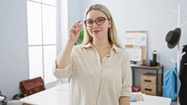 Gözlüklü, kendine güvenen genç, sarışın bir kadın modern işyerinde poz veriyor, profesyonellik ve tarz yayıyor..