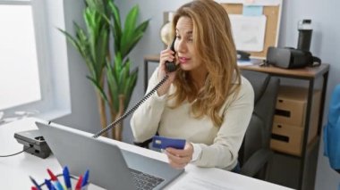 Ofiste birden fazla işe odaklanmış bir kadın, telefon ve dizüstü bilgisayarla, profesyonel dekorasyonun ortasında, kredi kartıyla uğraşıyor..