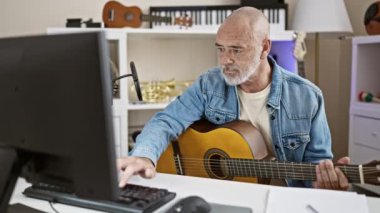 Evde çalışırken gitar çalan kot pantolonlu, kel, olgun bir adam.