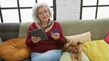 Kahverengi köpeğinin yanında tablet ve kredi kartı kullanan gözlüklü olgun bir kadın..