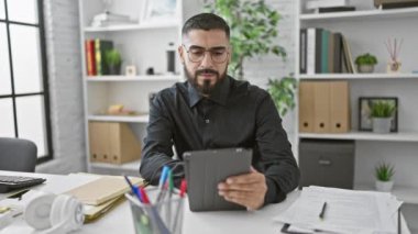 Modern ofis ortamında tablet kullanan düşünceli sakallı bir adam. Evrak işleri ve ofis malzemeleriyle çevrili..