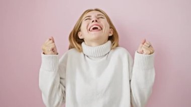 Heyecanlı sarışın güzellik zaferini kutluyor! Gözler kapalı, kollar havada, pembe ve izole arka planda muzaffer bir şekilde ayakta. Neşeli gülüşünü? saf zafer!