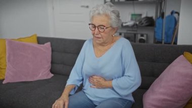 Gri saçlı, mavi kazaklı bir kadın evdeki gri bir kanepede otururken karın ağrısı çekiyor.
