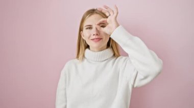 Neşeli, güzel sarışın kız parmaklarıyla ok işareti yapıyor, el dürbününden dikizliyor. İzole edilmiş pembe arka planda durarak, en mutlu, en ışıltılı gülümsemeyi yayarak