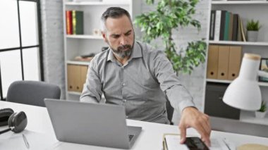 Modern bir ofiste çalışan, sakallı olgun İspanyol bir adam. Laptop ve akıllı telefon kullanıyor..