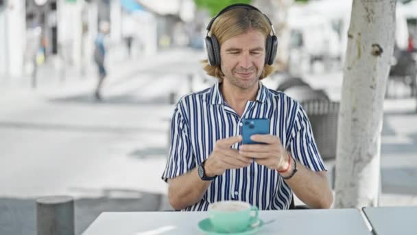 Мужчина Средних Лет Наслаждается Музыкой Наушниках Используя Смартфон Террасе Кафе — стоковое видео