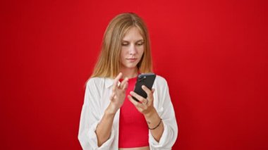 Heyecanlı, sarı saçlı genç bir kadın canlı kırmızı bir arka plana karşı akıllı telefon içeriğine tepki veriyor..