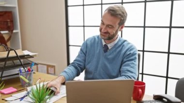 Gülümseyen orta yaşlı işadamı modern ofis ortamında akıllı telefon kullanıyor, bağlantı kuruyor..