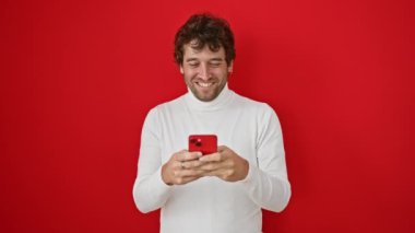 Sersemlemiş genç İspanyol bir adam, ağzı açık tamamen inançsız, akıllı telefon kullanırken yüzüne korku kazınmış, şaşırmış ama korkmuş, kırmızı arka planda soyutlanmış.