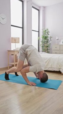 A young man practices advanced yoga poses on a blue mat in a bright, modern bedroom