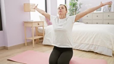 Günlük kıyafet giymiş genç bir kadın, düzenli yatak odasında pembe bir minder üzerinde yoga yapıyor, sükunet ve yaşam tarzı dengesini somutlaştırıyor..