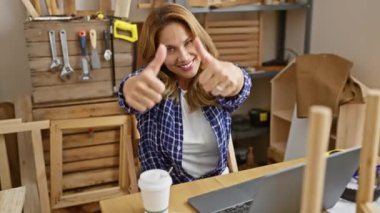 Marangozluk masasında oturan neşeli genç Latin kadın, gülümseyen ve parlayan kazananın başparmakları başarı ve mutlulukta.