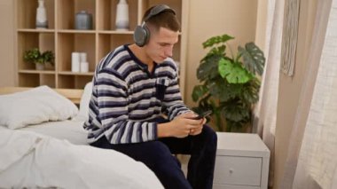 Rahatlamış kulaklıklı bir adam rahat bir yatak odasında bitkiler ve minimalist dekorlarla akıllı telefon kullanır.