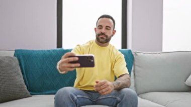 Sarı tişörtlü dövmeli adam, gri koltukta selfie çekiyor. İçeriden neşe ve yaşam tarzı gösteriyor..