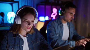 Odaklanmış kadın ve erkek, geceleri neon ışıklı bir oyun odasında video oyunu oynayarak heyecan ve konsantrasyonu ifade ediyorlar..