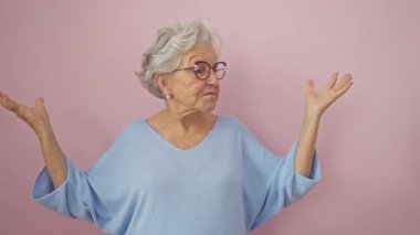 Şaşkın orta yaşlı kadın gözlük takıyor ve süveterin de hiçbir şeyden haberi yok. Gri saçlı yaşlı kadın şaşkın duruyor, pembe izole edilmiş arka plan için omuz silkiyor.