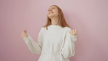 Heyecanlı genç kızıl saçlı kadın zafer duruşu içinde, neşeyle gülümsüyor, kazak giyiyor, galibiyetini haykırıyor, pembe arka planda izole edilmiş.