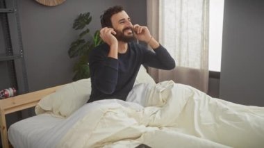Kulaklıklı, gülümseyen sakallı adam güneşli yatak odasında müzik dinliyor.