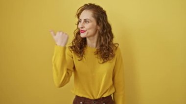 Neşeli genç bir kadın güvenli bir şekilde baş parmağını açık ağızlı bir gülümseme ile işaret ederek izole edilmiş sarı bir arka plana karşı duruyor. Olumlu ve anlamlı duygularla dolu bir portre..