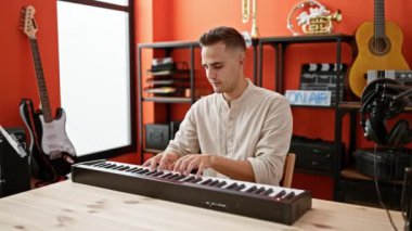 Odaklanmış genç bir adam, gitarlar ve kayıt ekipmanlarıyla çevrili canlı bir müzik stüdyosunda klavye çalıyor..