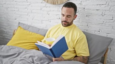 Yakışıklı dövmeli adam evde kahve fincanıyla yatakta kitap okuyor.