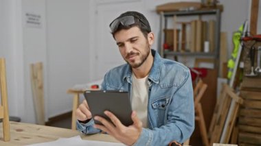 Sakallı düşünceli genç bir adam marangozluk atölyesindeki bir tabletin planlarını gözden geçiriyor..
