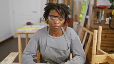 Rastalı genç siyahi kadın marangozluğun zaferini kutluyor, güvenlik gözlüklerini takarken yüzünde zaferin coşkulu ifadesi, neşeli bir gülümsemeyle zaferini kutluyor.