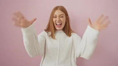 Genç, heyecanlı kızıl saçlı kadın, başarının sevinciyle, zaferini kutluyor, ayakta duruyor, kollarını zaferle kaldırıyor, izole edilmiş pembe bir duvara karşı kazak giyiyor..