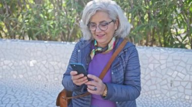 Arka planda yeşillik olan bir parkta akıllı telefon kullanan orta yaşlı bir kadın..