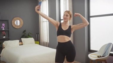 Genç bir kadın şık döşeli odasında selfie çekerken kaslarını esnetiyor.