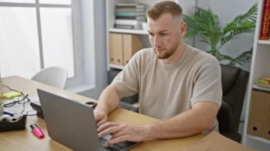 Genç sakallı bir adam, modern bir ofiste dizüstü bilgisayarda çalışıyor. Rahatsızlık ve göğüs ağrısını sağlık konseptiyle ifade ediyor..