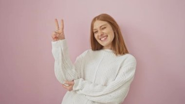 Kendine güvenen genç kızıl saçlı kadın göz kırparak gülümsüyor, süveterini sallıyor ve pembe izole bir arka planda barış işareti gösteriyor.