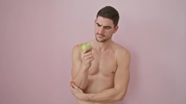 Kollarında elmayla tişörtsüz, cahil İspanyol genç adam, soyutlanmış pembe arka planda 'hiçbir fikrim' ifadesini sergiliyor..