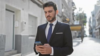 İyi giyimli, sakallı, güneşli bir caddede yürürken akıllı telefon tebessümleri kullanan İspanyol bir adam..