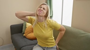 Genç sarışın bayan, ağrılı boyun ağrısı yüzünden acı çekiyor, evinde yaralanmış, acı çekerken tişört giyiyor.