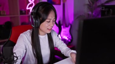 Geceleri karanlık bir oyun odasında kulaklık takan endişeli Asyalı kadın bilgisayar kullanıyor.