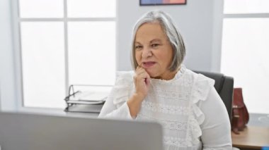 Gri saçlı düşünceli bir son sınıf öğrencisi parlak ofis ortamında dizüstü bilgisayar kullanırken nazikçe gülümsüyor..