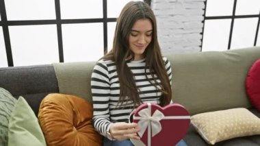 Hispanik genç, elinde kalp şeklinde bir hediye kutusu tutarken gülümsüyor, kanepede oturuyor..