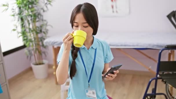 Młoda Azjatycka Kobieta Pije Kubka Podczas Sprawdzania Smartfona Klinice — Wideo stockowe