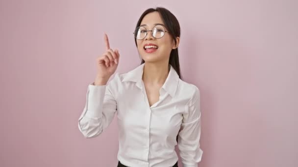 兴奋的中国年轻女子 戴着眼镜 自信地站在那里 快乐地微笑着 她的手指指向一个漂亮而孤立的粉色背景 带着一个聪明的想法 — 图库视频影像