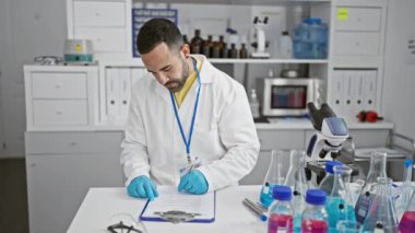 Sakallı İspanyol bir adam laboratuarda yazıyor. Eldiven takmış, labirent giymiş, bilimsel ekipmanlarla çevrili..