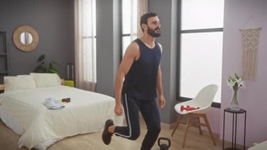 Sakallı adam modern bir yatak odasında egzersiz yapıyor, kolsuz tişört ve eşofmanla hareket ediyor..