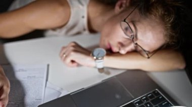 Ofis masasında uyuyan gözlüklü yorgun bir kadın, loş bir odada dizüstü bilgisayar, saat ve kağıtlar gösteriyor..