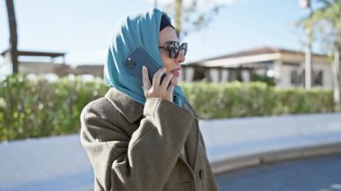 Açık havada güneşli bir günde tesettürlü bir kadın güneş gözlüğü ve palto giyerken akıllı telefonuyla konuşur..