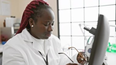 Gözlüklü ve laboratuvar önlüklü gülümseyen Afrikalı bir kadın araştırma laboratuarında çalışıyor..