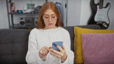 Beyaz kazaklı beyaz bir kadın gri bir koltukta akıllı telefon kullanıyor. Kapalı bir yerde. Arka planda gitar var..