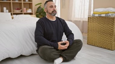 A mature man with a beard meditates cross-legged in a serene bedroom setting, depicting tranquility