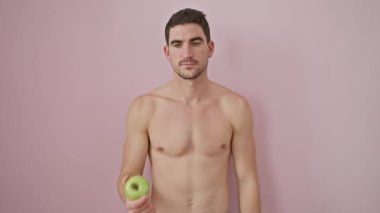 Gömleksiz, genç, İspanyol bir adam, neşeli bir şekilde, sağlıklı bir elma sunuyor. Pembe izole bir arka planda kamera için kendinden emin ve mutlu görünüyor.
